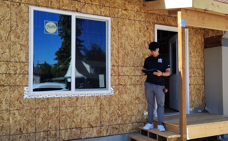 An H Design team member wearing a branded company shirt while reviewing code compliance and building permit documentation on a digital tablet at an active residential home construction site.